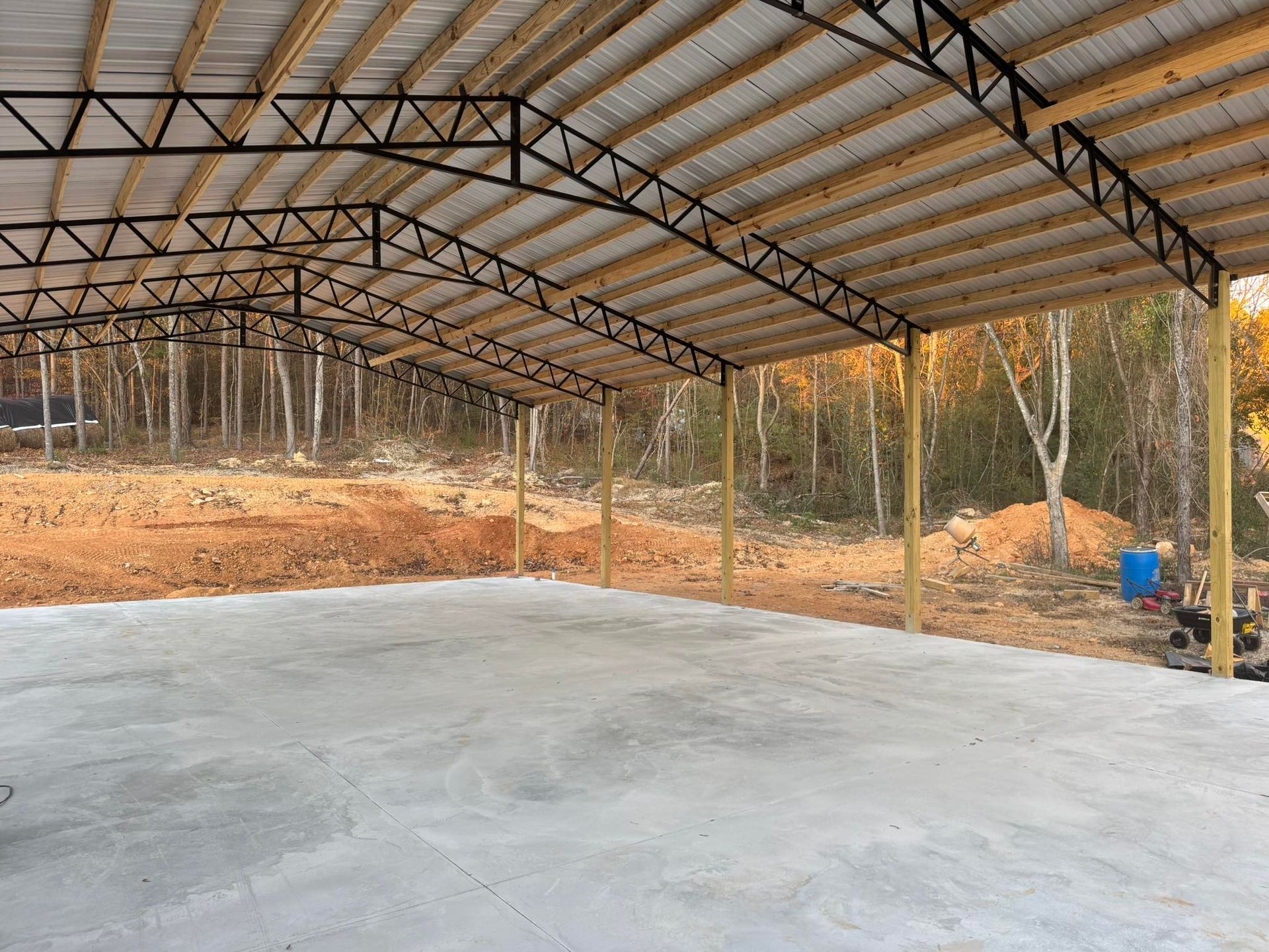 Open-air metal and wood structure on a concrete pad with a treeline background.
