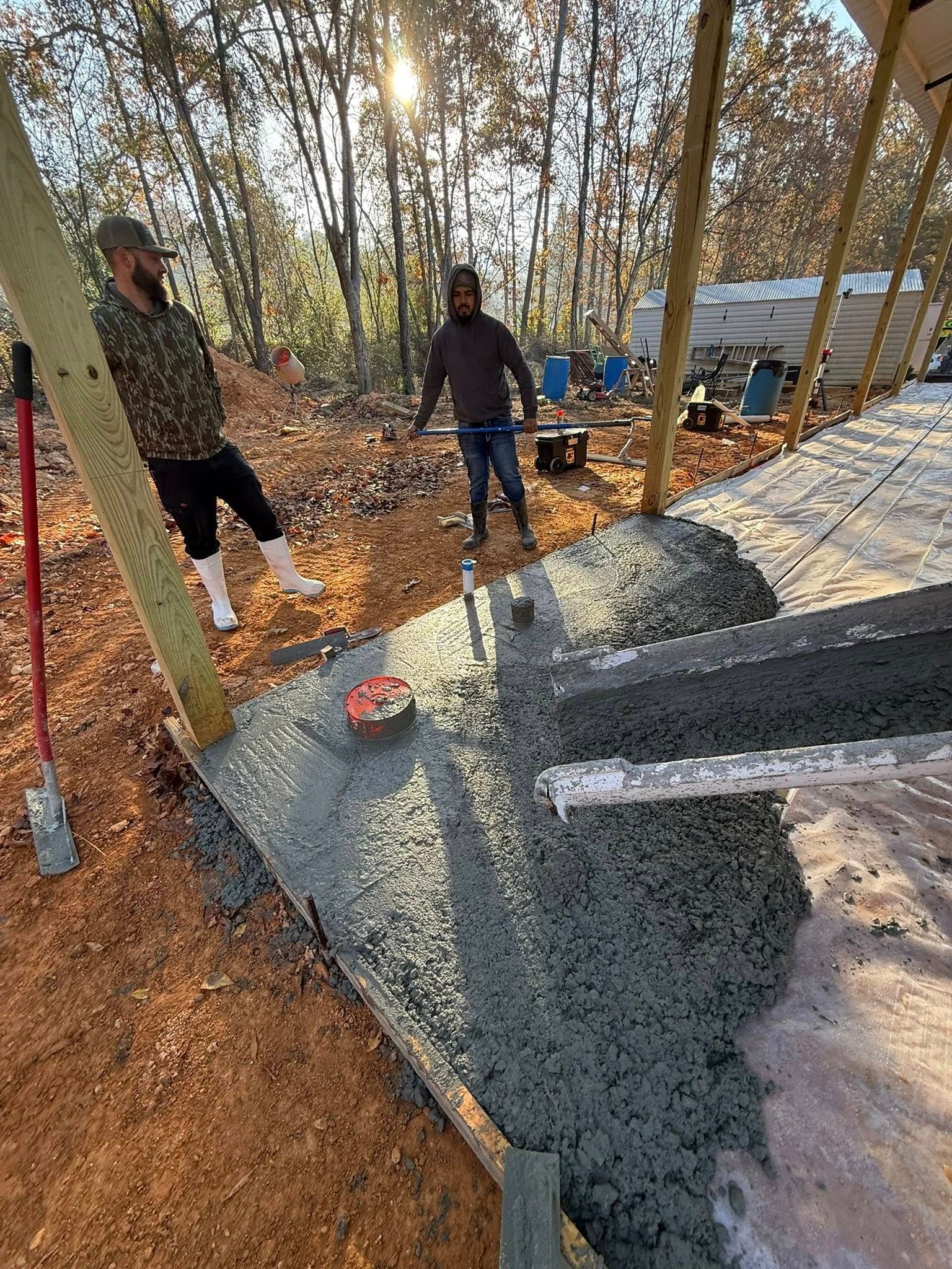 Two people pouring concrete for a foundation, using tools in an outdoor wooded setting.