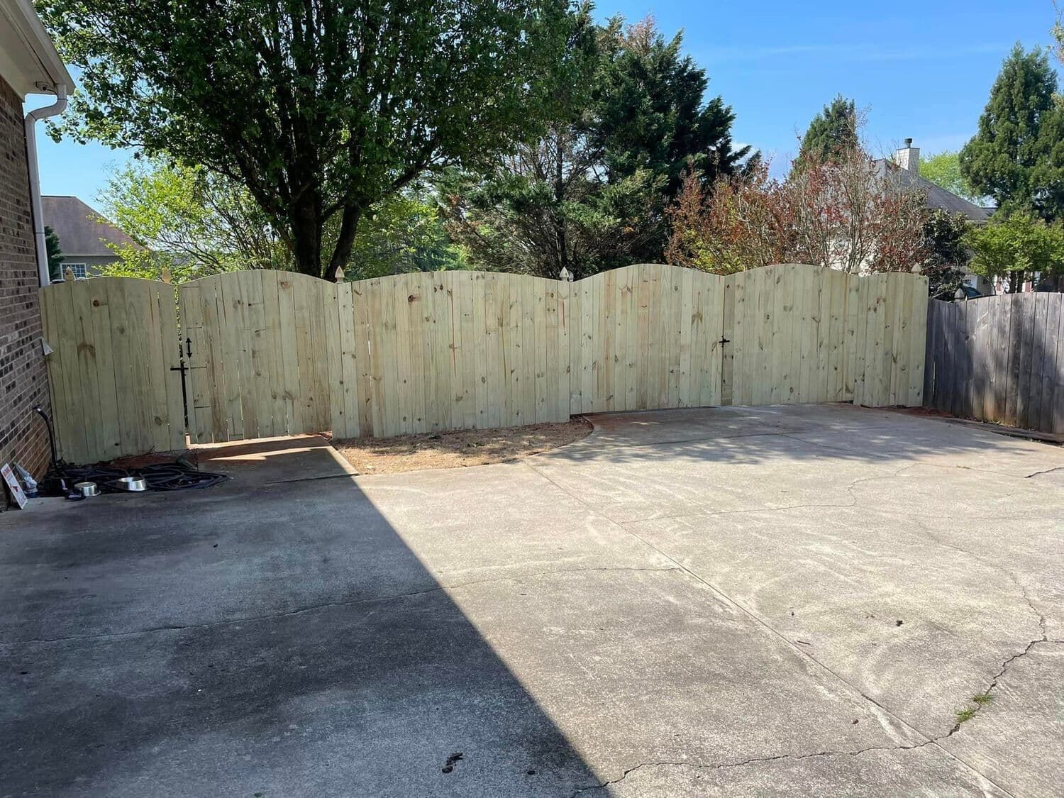 Wooden fence in backyard, sunny day. Concrete area in foreground. Trees in background.