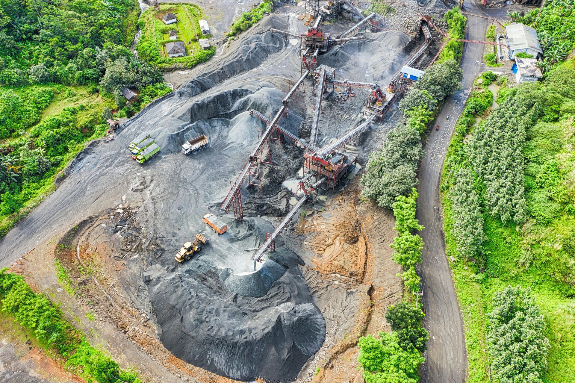 Aerial view of a quarry site with machinery, trucks, and piles of gravel, surrounded by green trees.