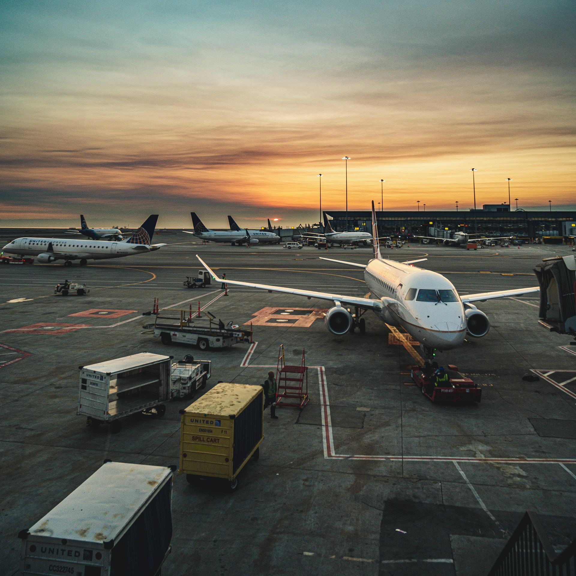 Airplanes on tarmac at sunset. Several planes, ground crew, cargo containers, airport terminal, orange and blue sky.
