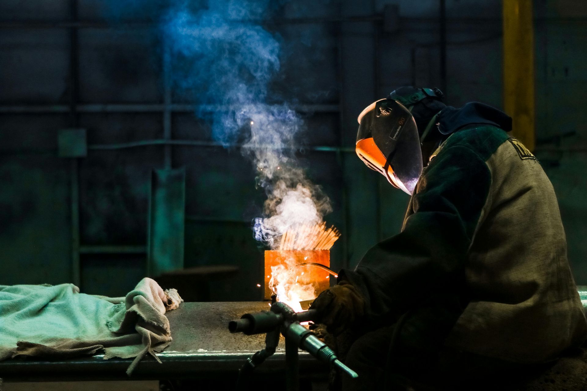 Welder in protective gear working with a torch, creating sparks and smoke in a workshop.