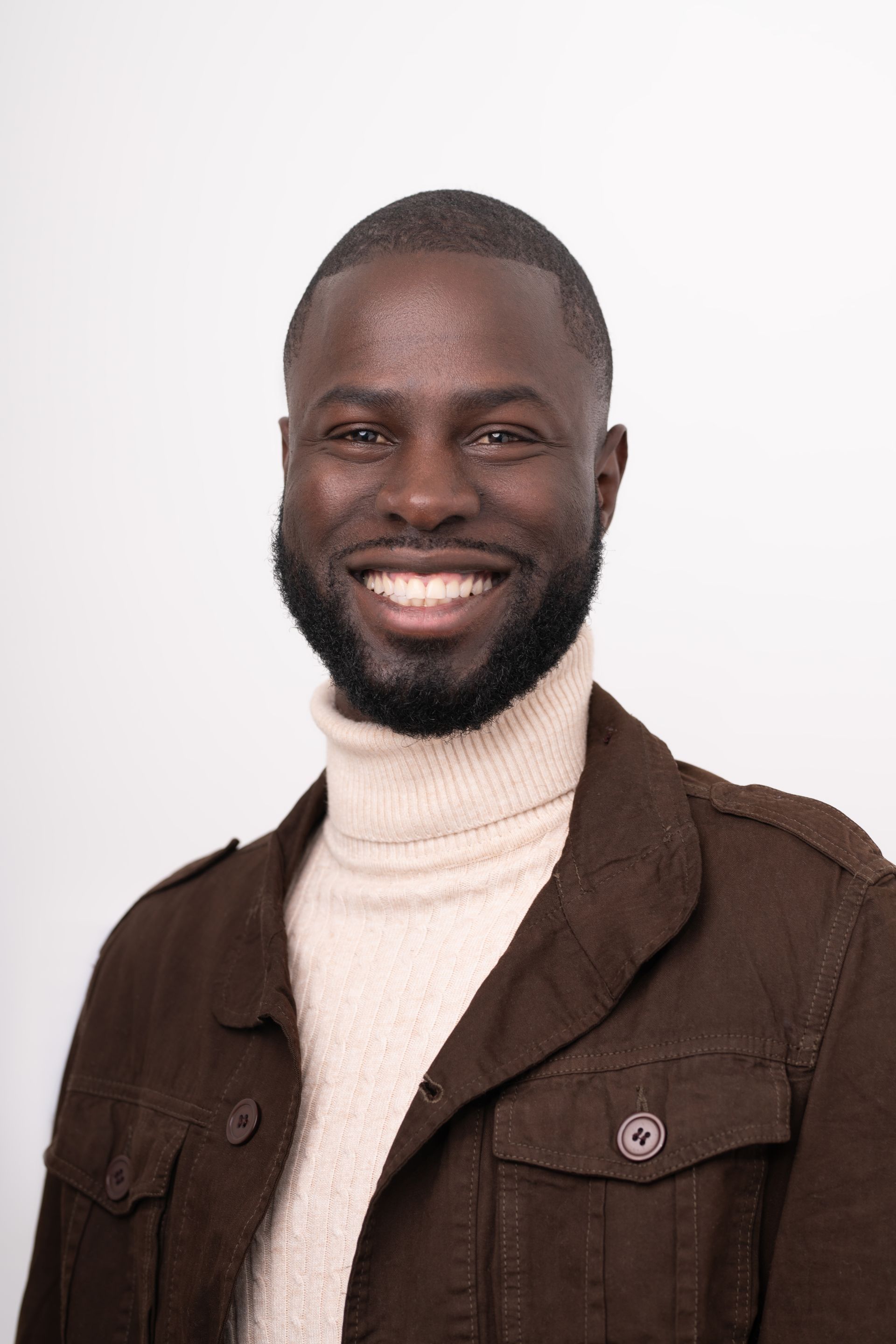 Un homme avec une barbe porte une veste marron et un col roulé blanc.