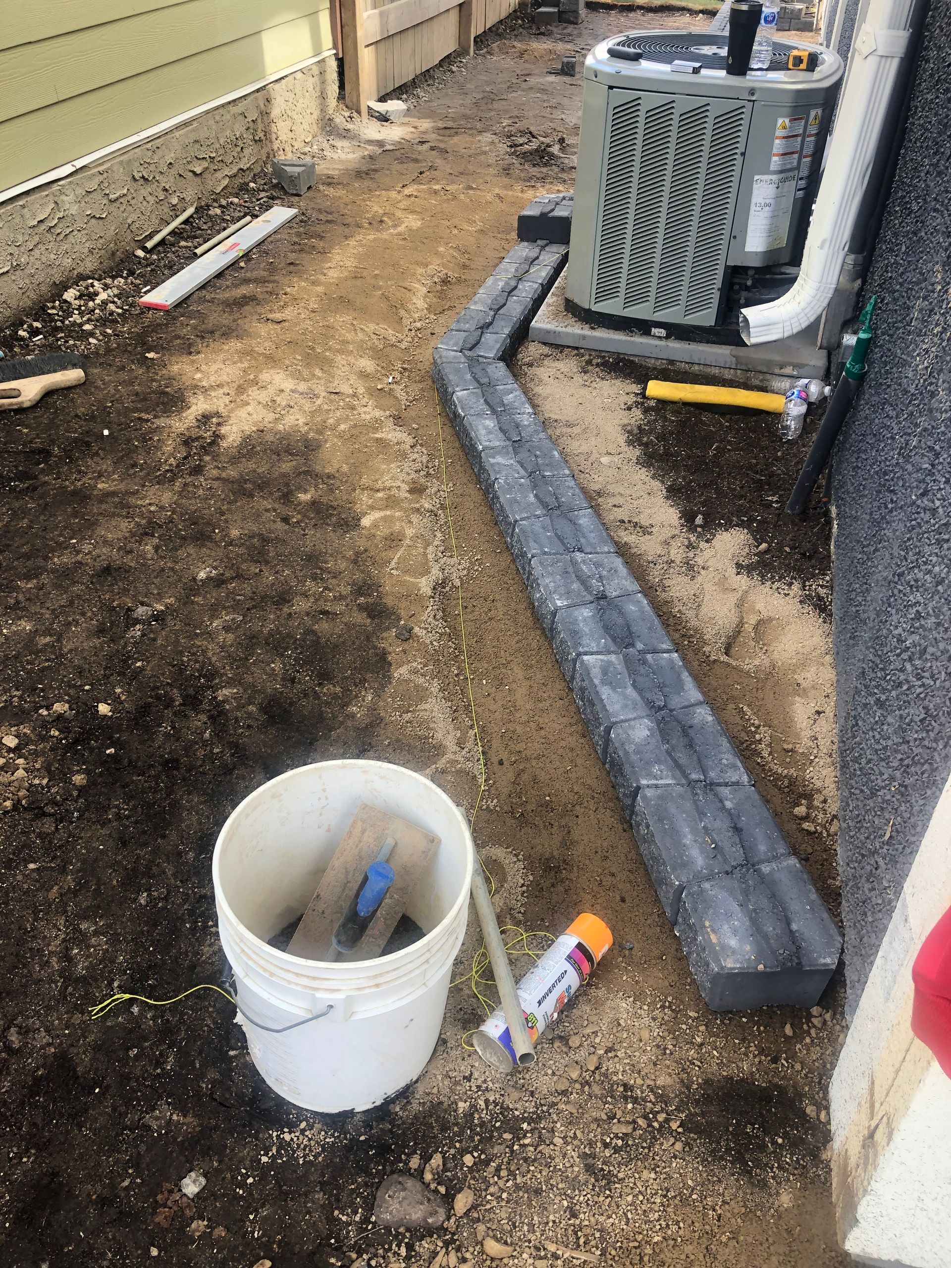 A bucket of tools is sitting on the ground next to an air conditioner.