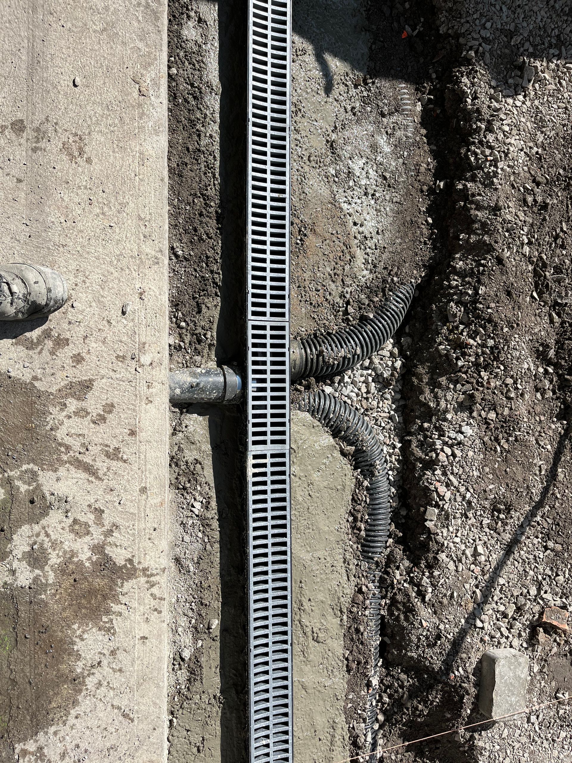 A metal drain is sitting in the dirt next to a concrete wall.