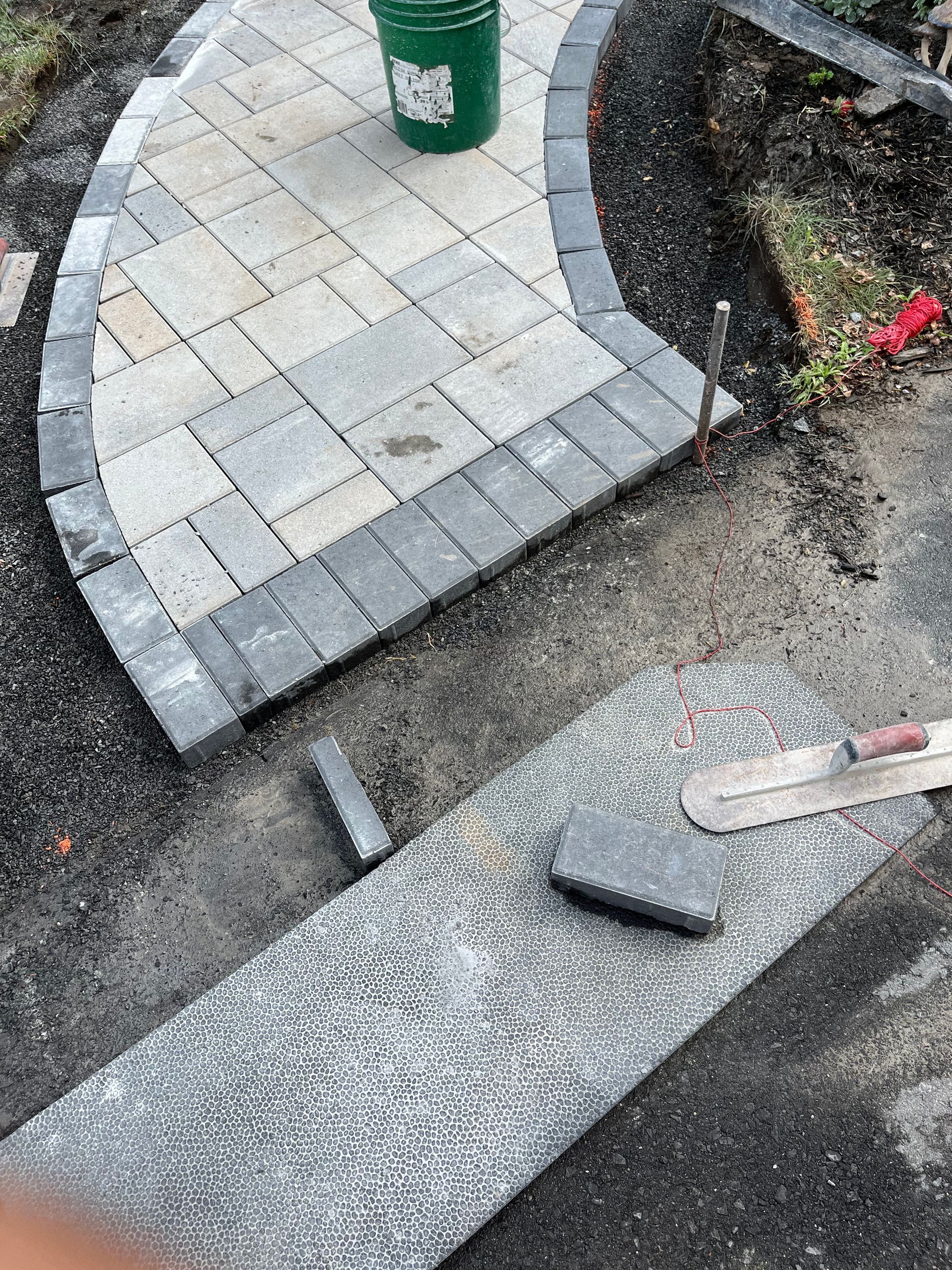 A person is laying bricks on a sidewalk next to a green bucket.