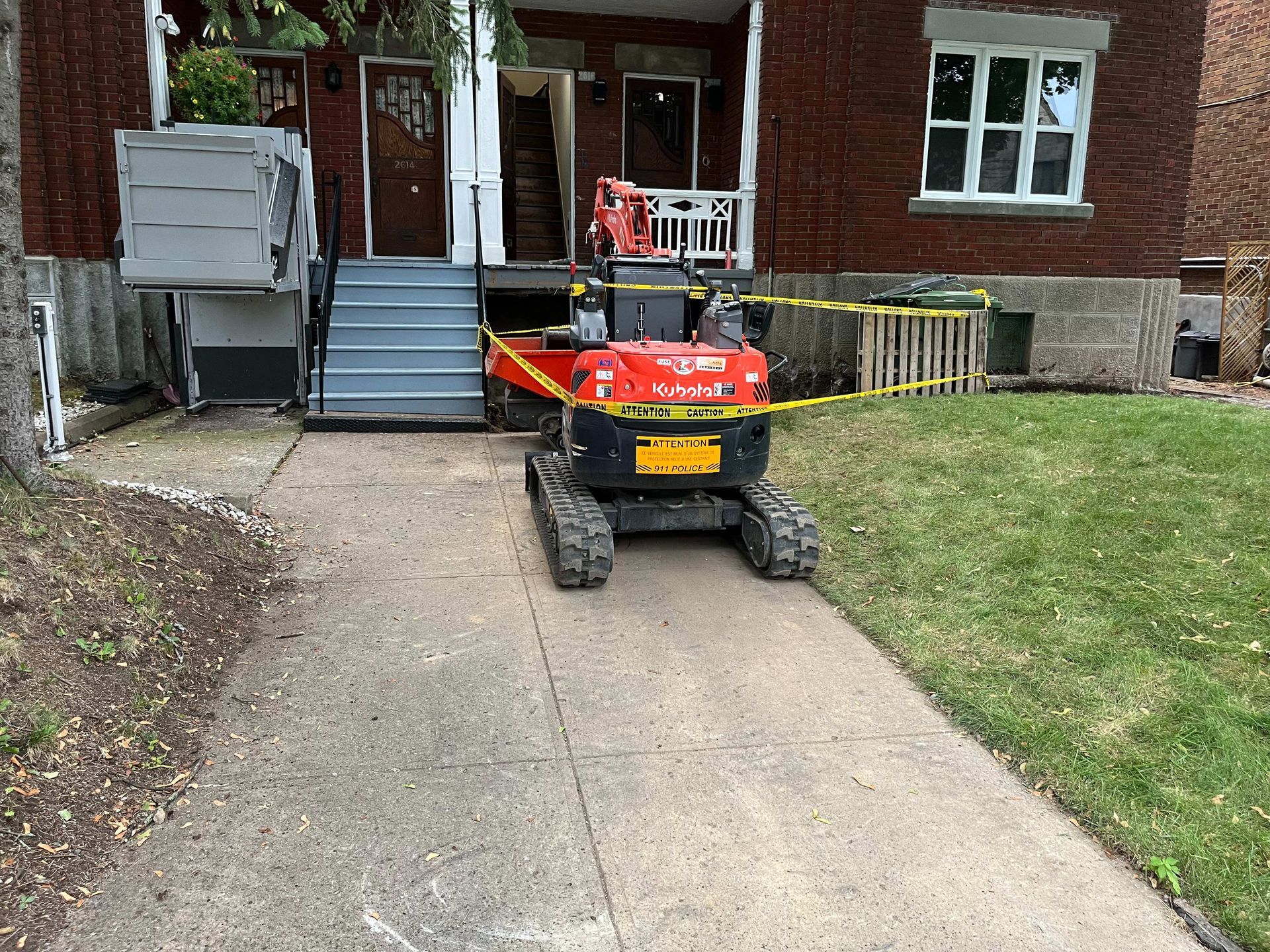 A small red excavator is parked on the sidewalk in front of a brick house.