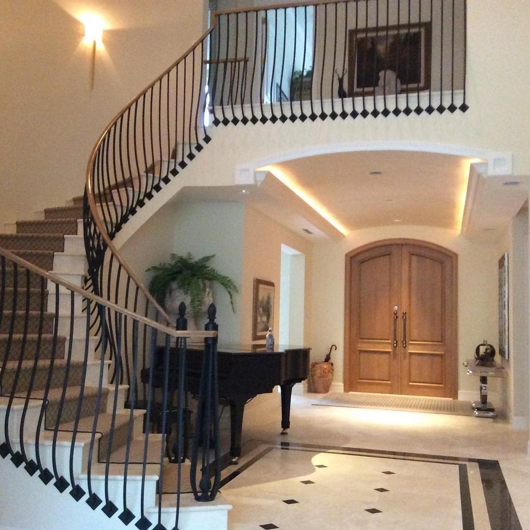 Elegant foyer with curved staircase, piano, and double doors; light wood and marble floors.