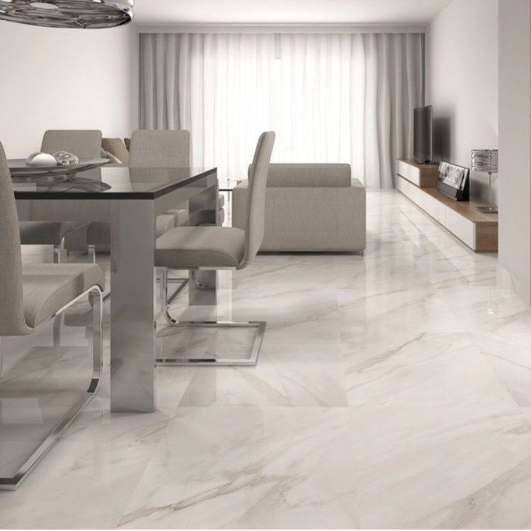 Dining room with a glass table, chrome chairs, and marble-like flooring. A white sofa and TV are in the background.