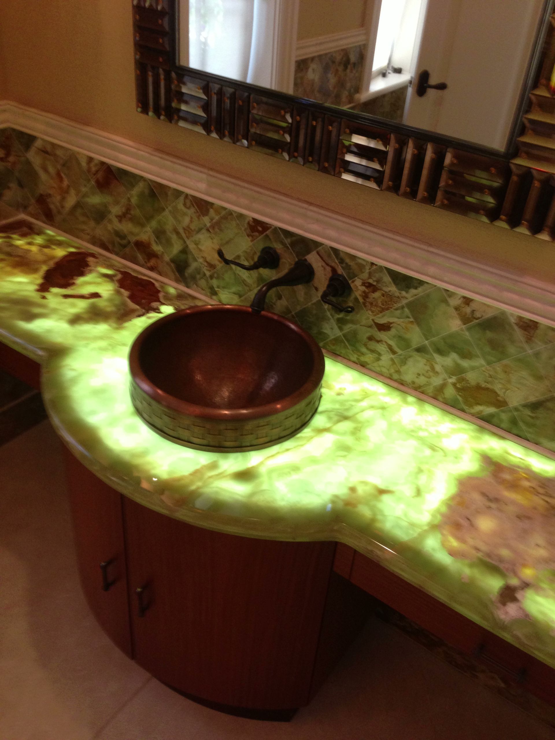 Bathroom vanity with a backlit green countertop, copper sink, and decorative mirror.
