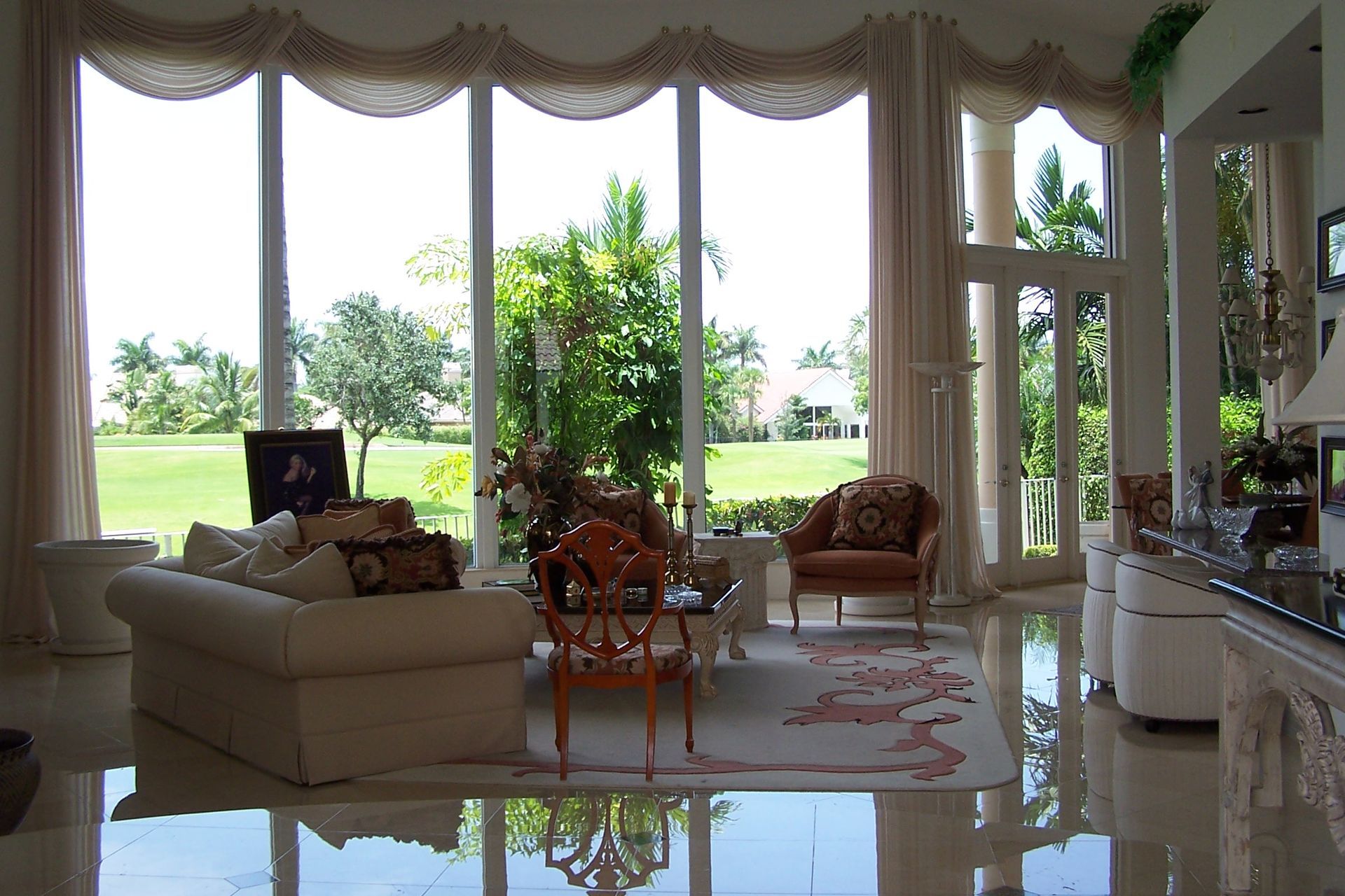 Living room with large windows overlooking a green lawn. Beige furniture, ornate rug, and curtains.