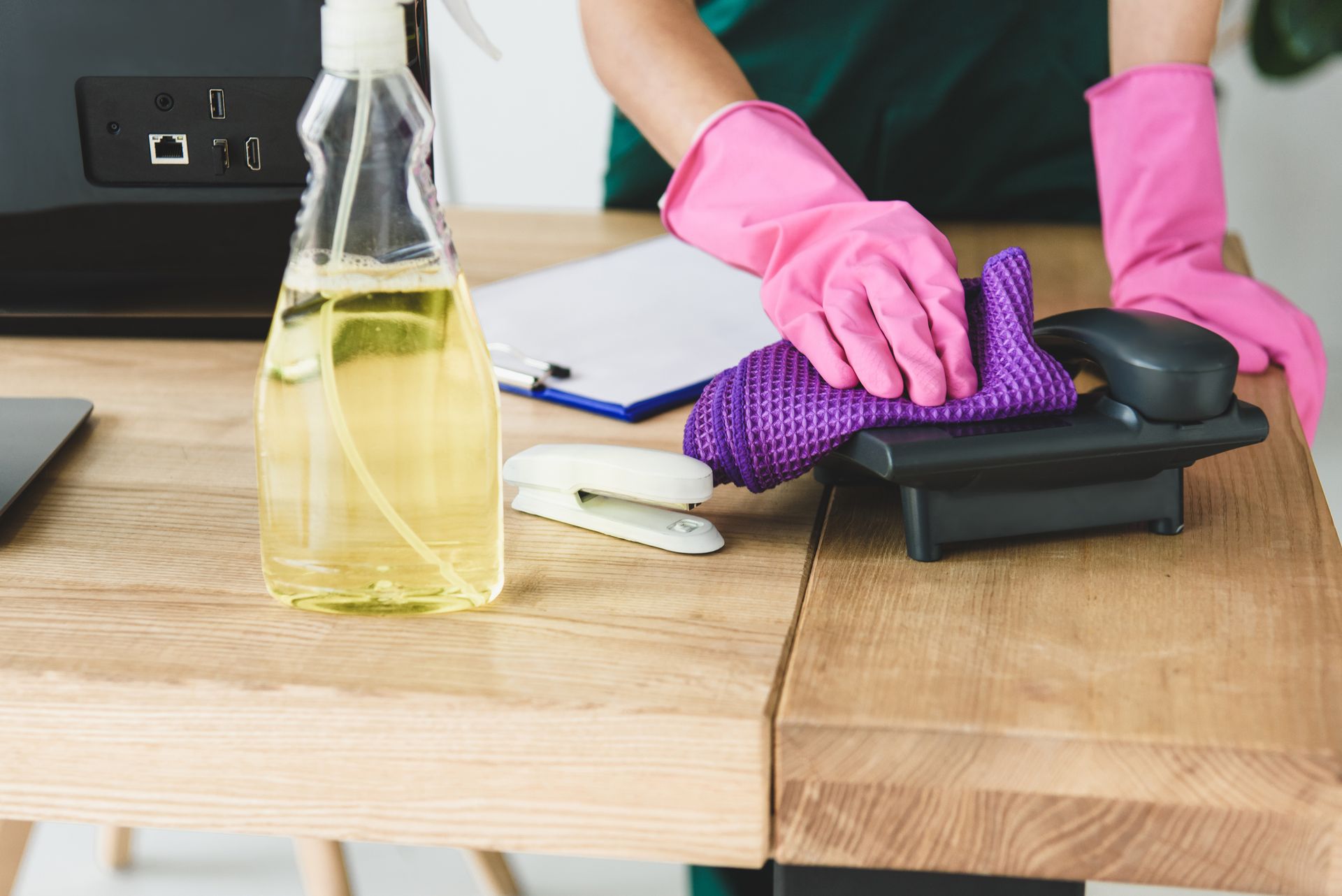 Person wearing pink gloves cleaning a wooden desk with a purple cloth and spray bottle.