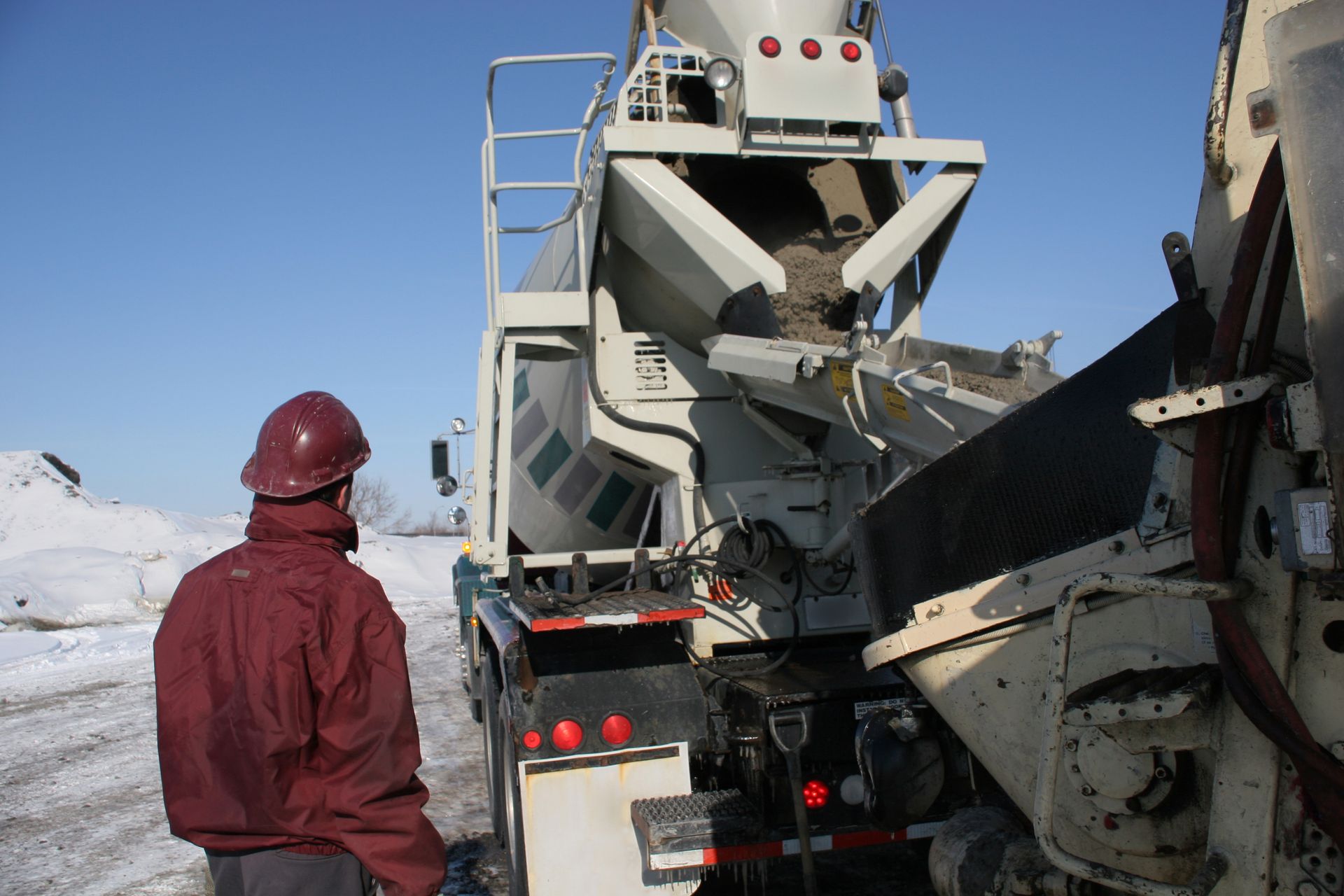 Person in maroon jacket and hard hat watches concrete pour from a mixer truck on a snowy day.