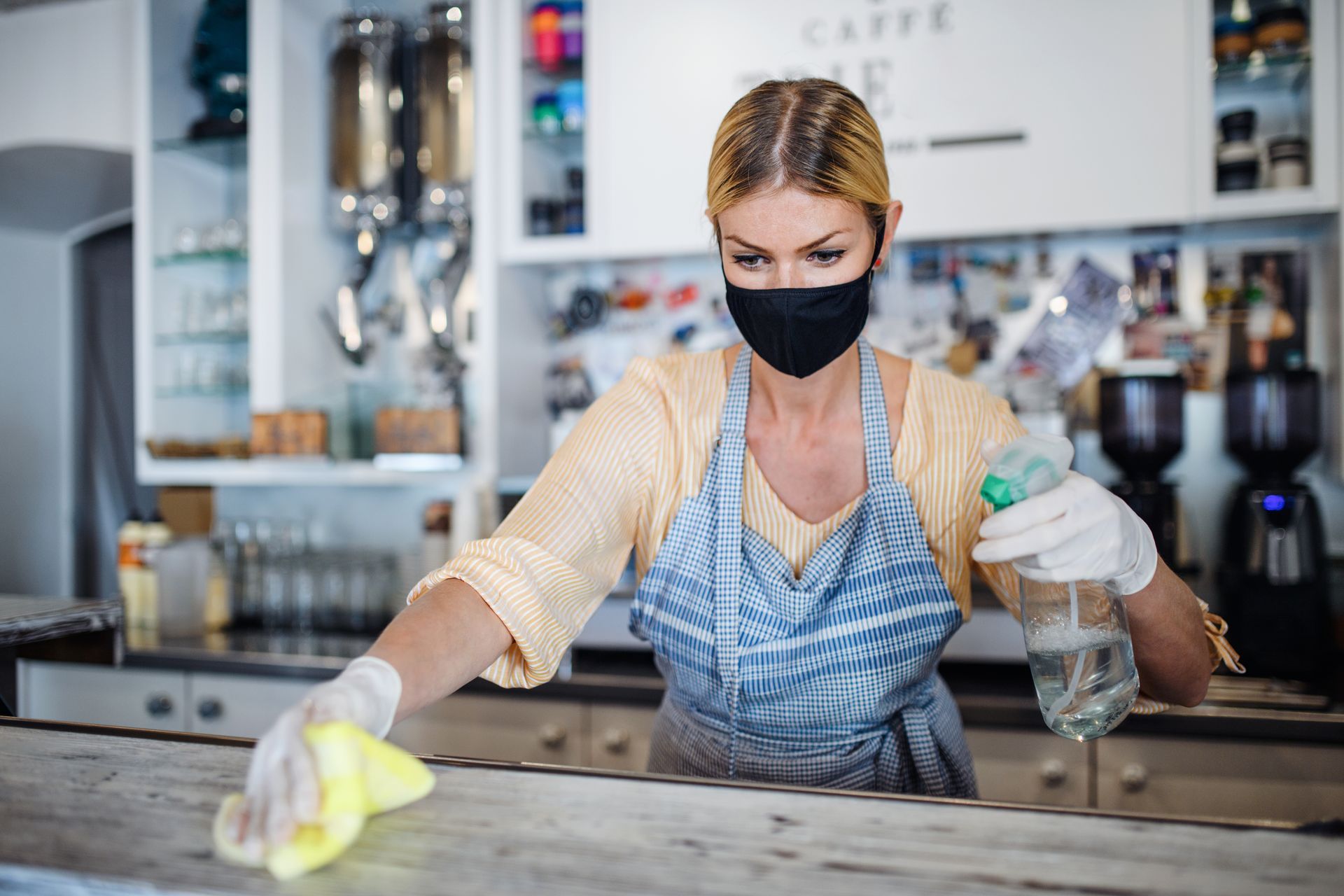 A woman wearing a mask and gloves is cleaning a counter in a restaurant.