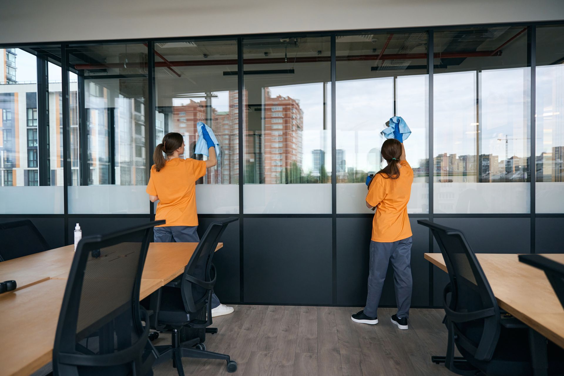 Two people wearing orange shirts and dark pants clean a large glass office partition with blue cloths.