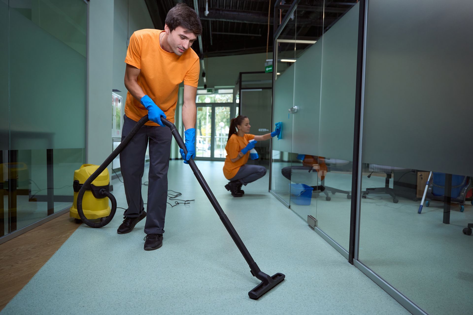 Two people cleaning an office. One vacuums floor, the other wipes glass wall. Both wear orange shirts, blue gloves.