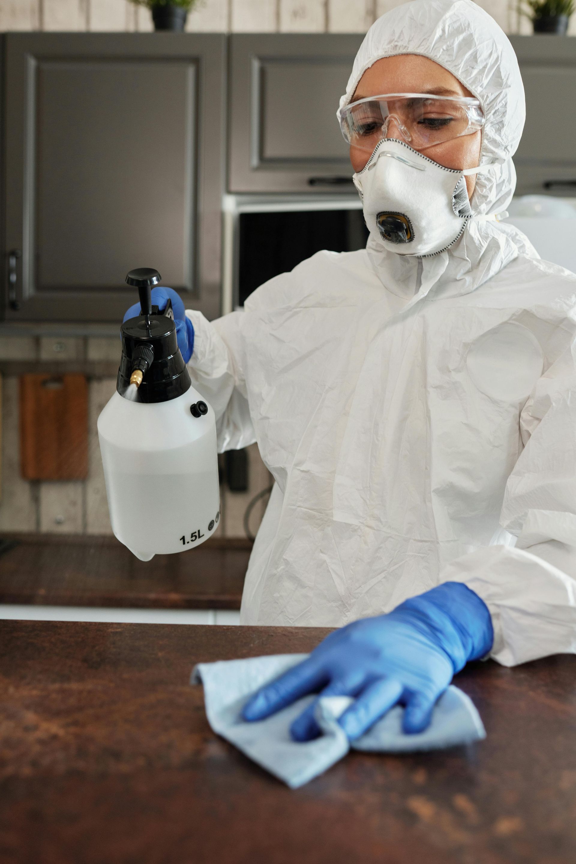 A woman in a protective suit is cleaning a counter with a spray bottle.