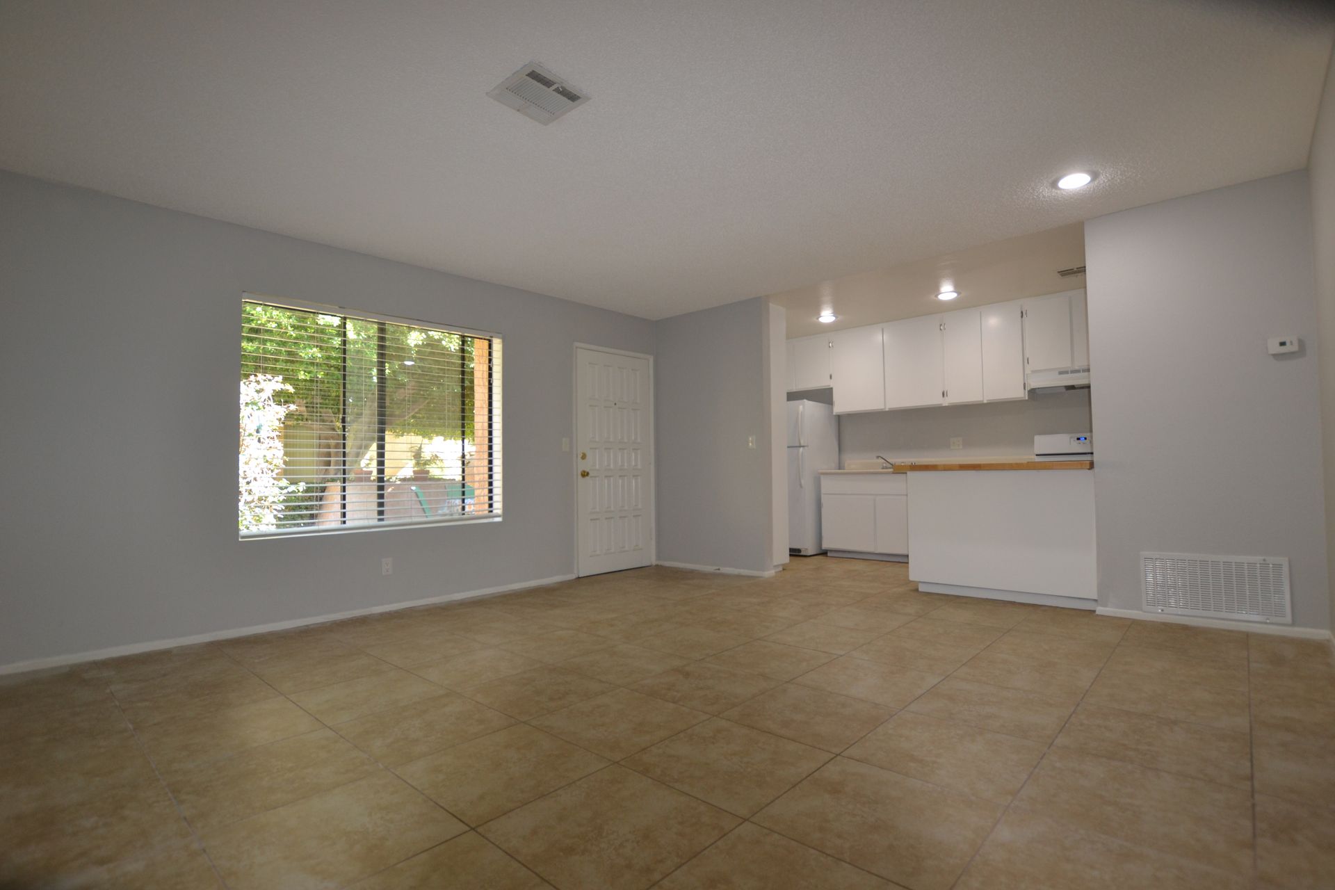An empty living room with a large window and a kitchen in the background.