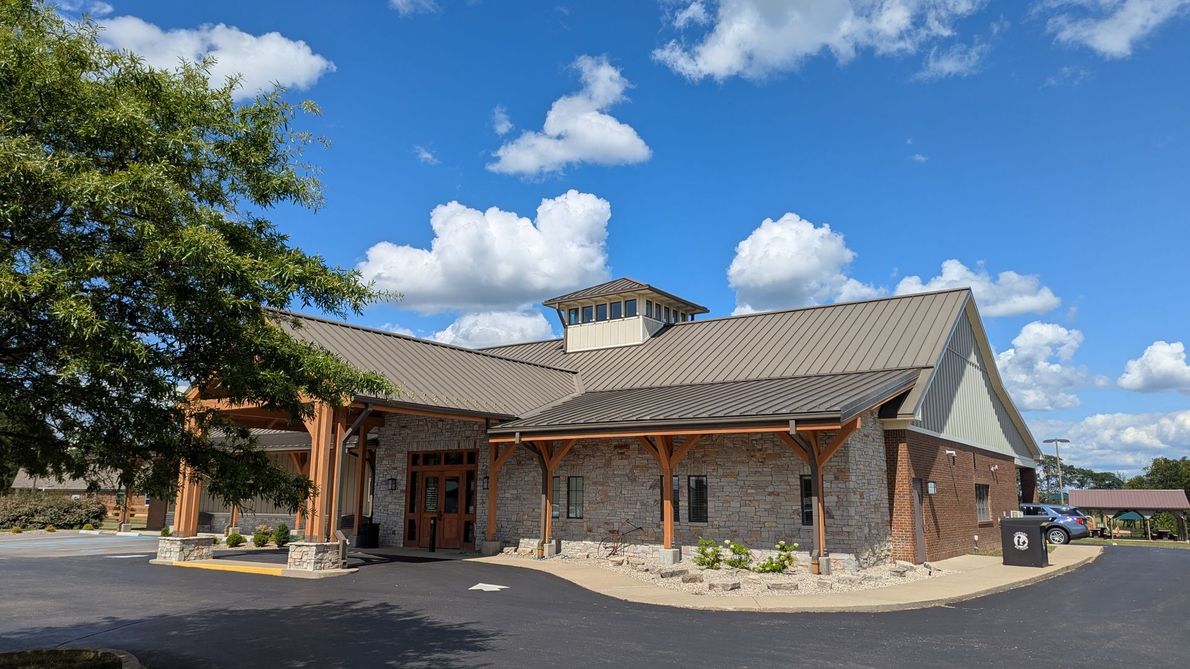 Brick building with a gray roof beside a tree-lined parking lot under a blue sky