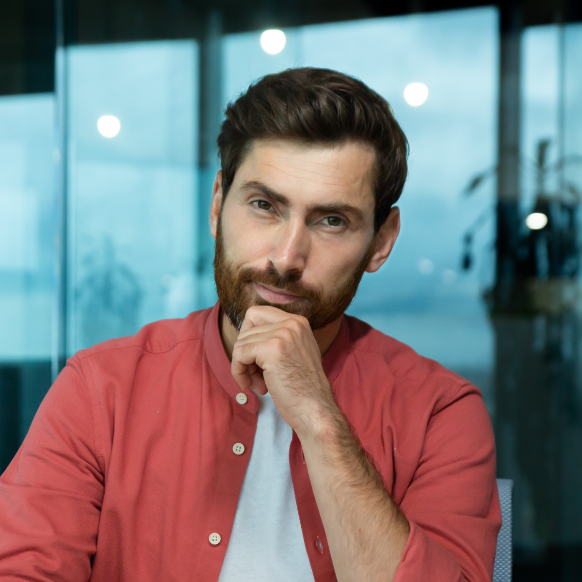 A man with a beard is wearing a red shirt and has his hand on his chin.