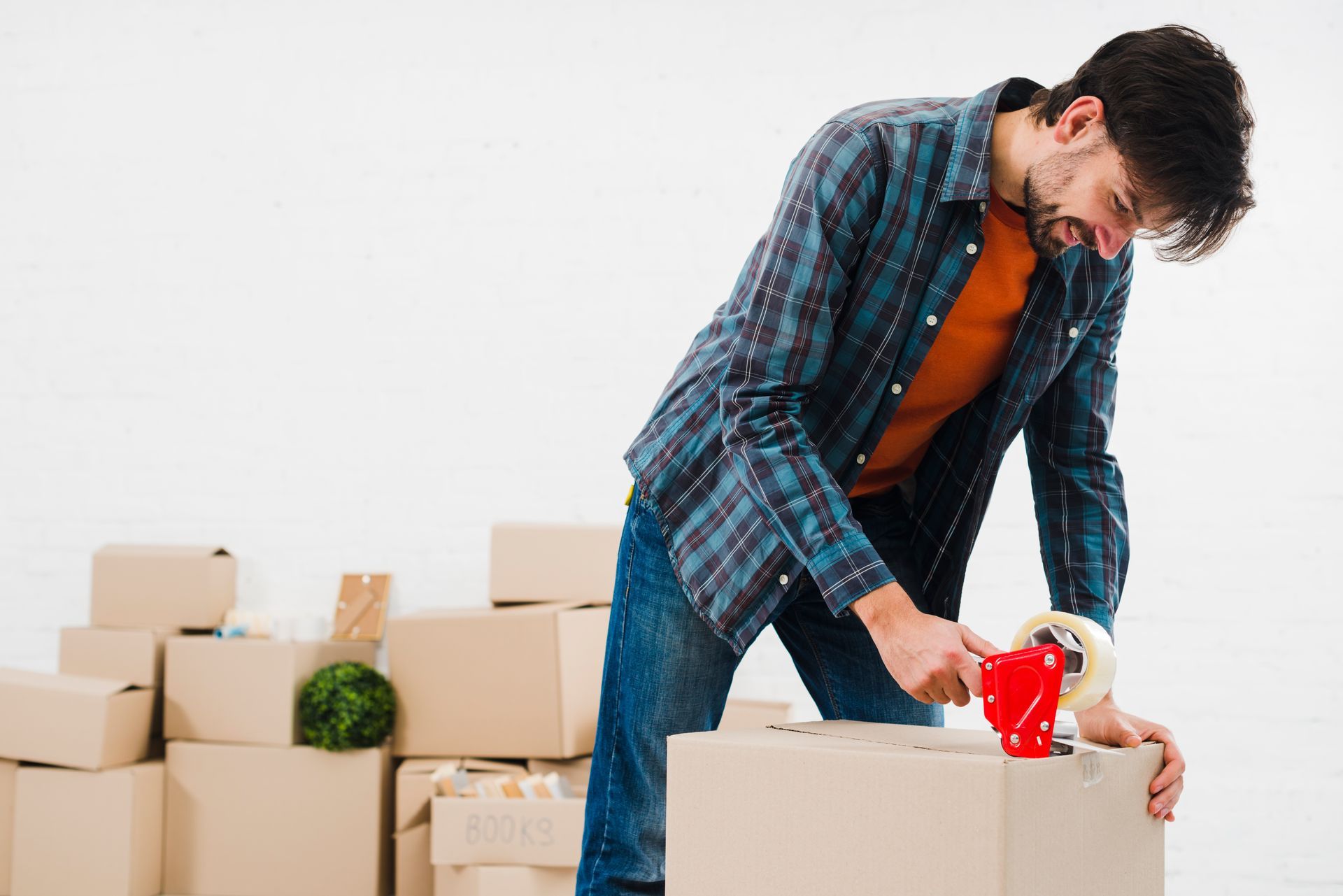 Man taping cardboard box while moving, surrounded by more boxes. White backdrop.
