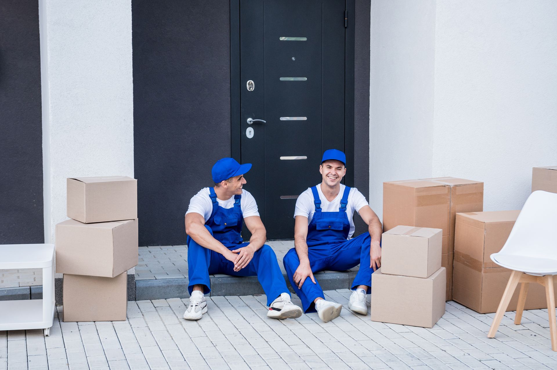 Two movers are sitting on the steps of a house surrounded by cardboard boxes.