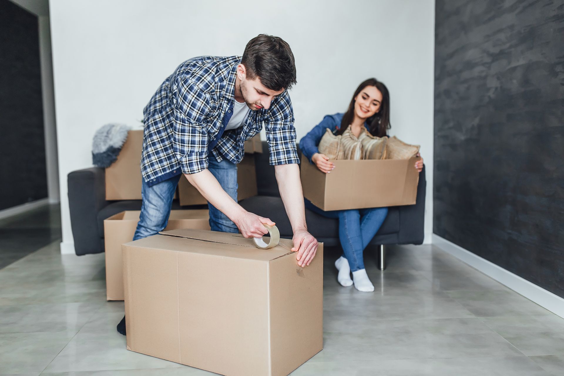 Man taping a moving box, woman packing another box on a couch, indoors.