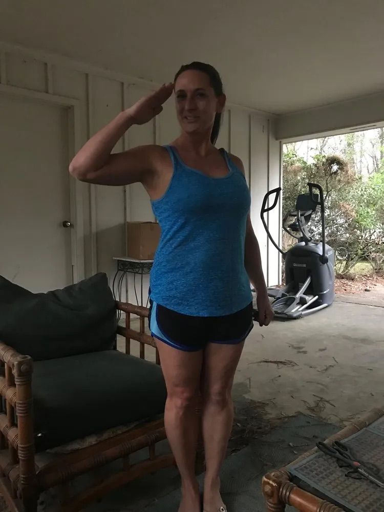 A woman in a blue tank top and shorts is saluting in a garage.