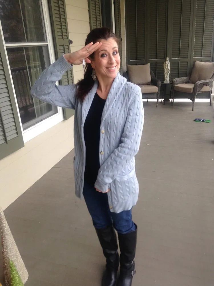 A woman wearing a light blue cardigan and black boots is standing on a porch.