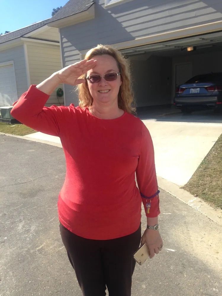 A woman in a red shirt salutes in front of a garage