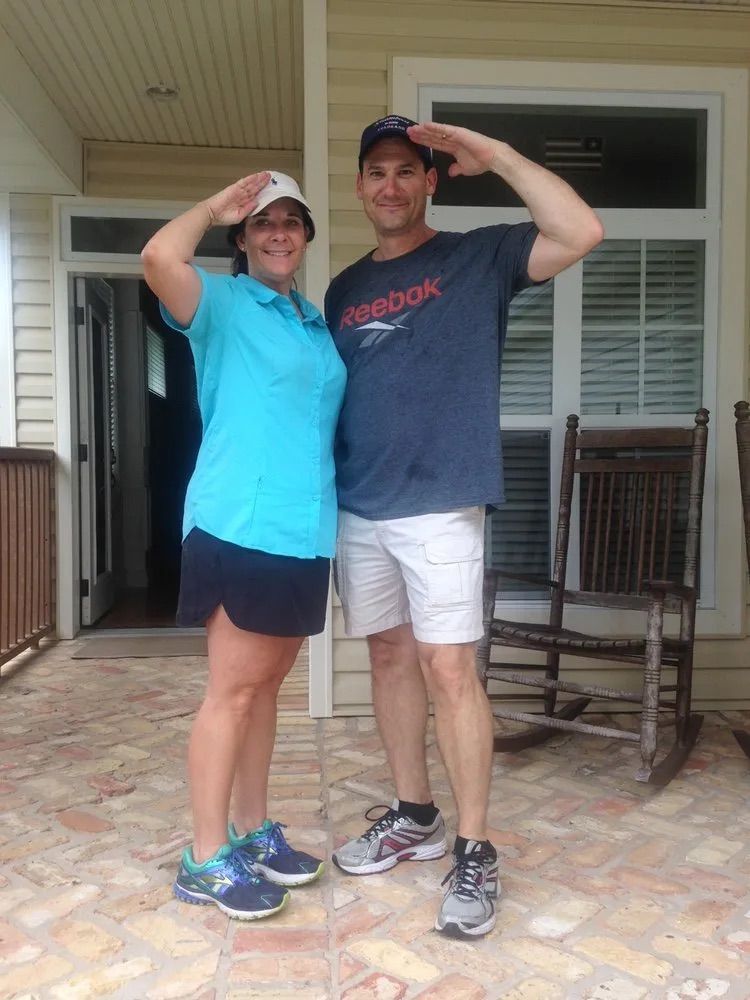 A man and a woman are posing for a picture on a porch . the man is wearing a reebok shirt.