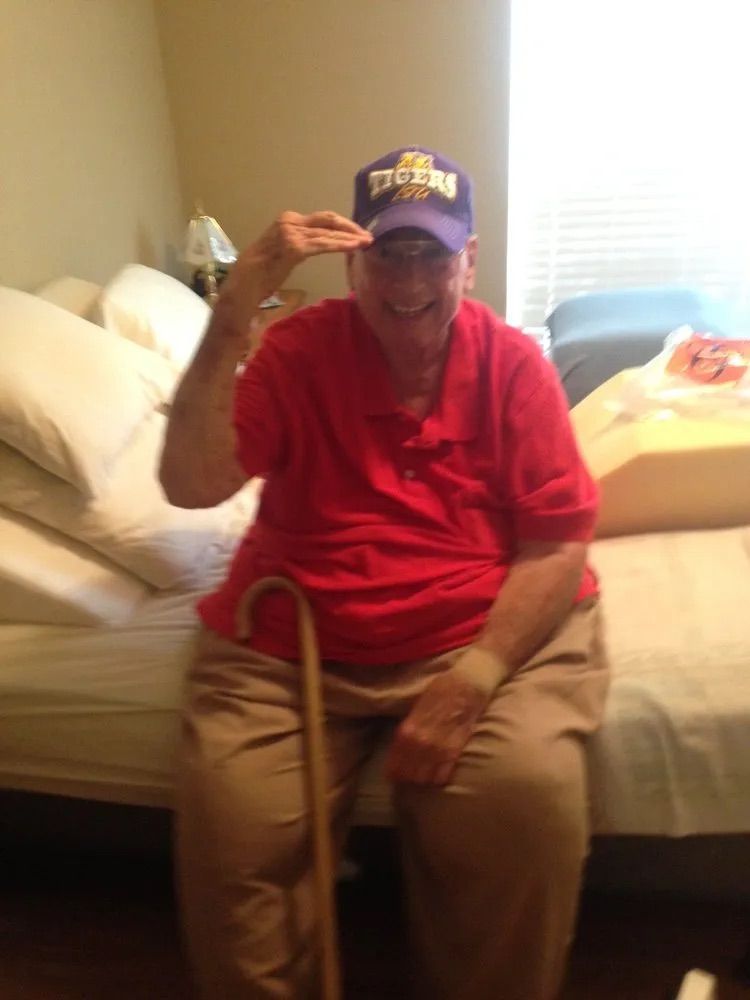A man wearing a red shirt and a tigers hat is sitting on a bed with a cane.