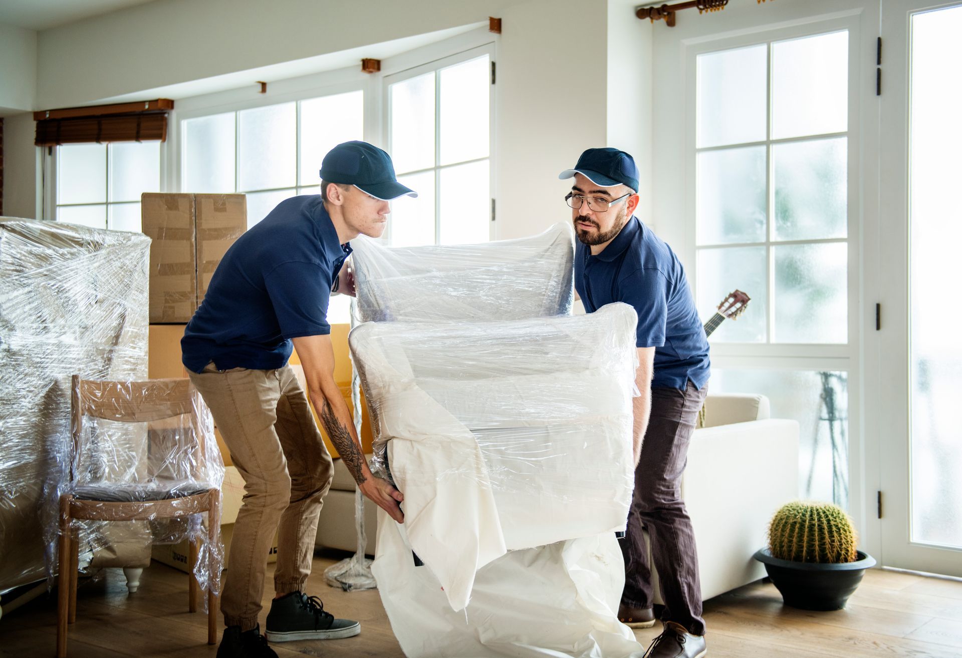 Two men are carrying a couch wrapped in plastic in a living room.