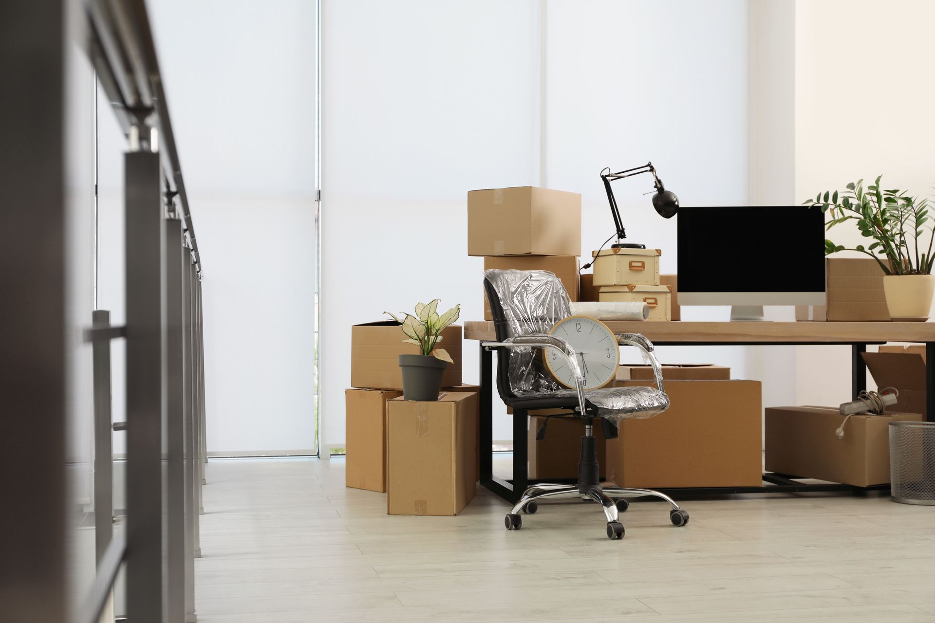 Office with moving boxes and desk, chair covered, plants, and computer, near a railing.