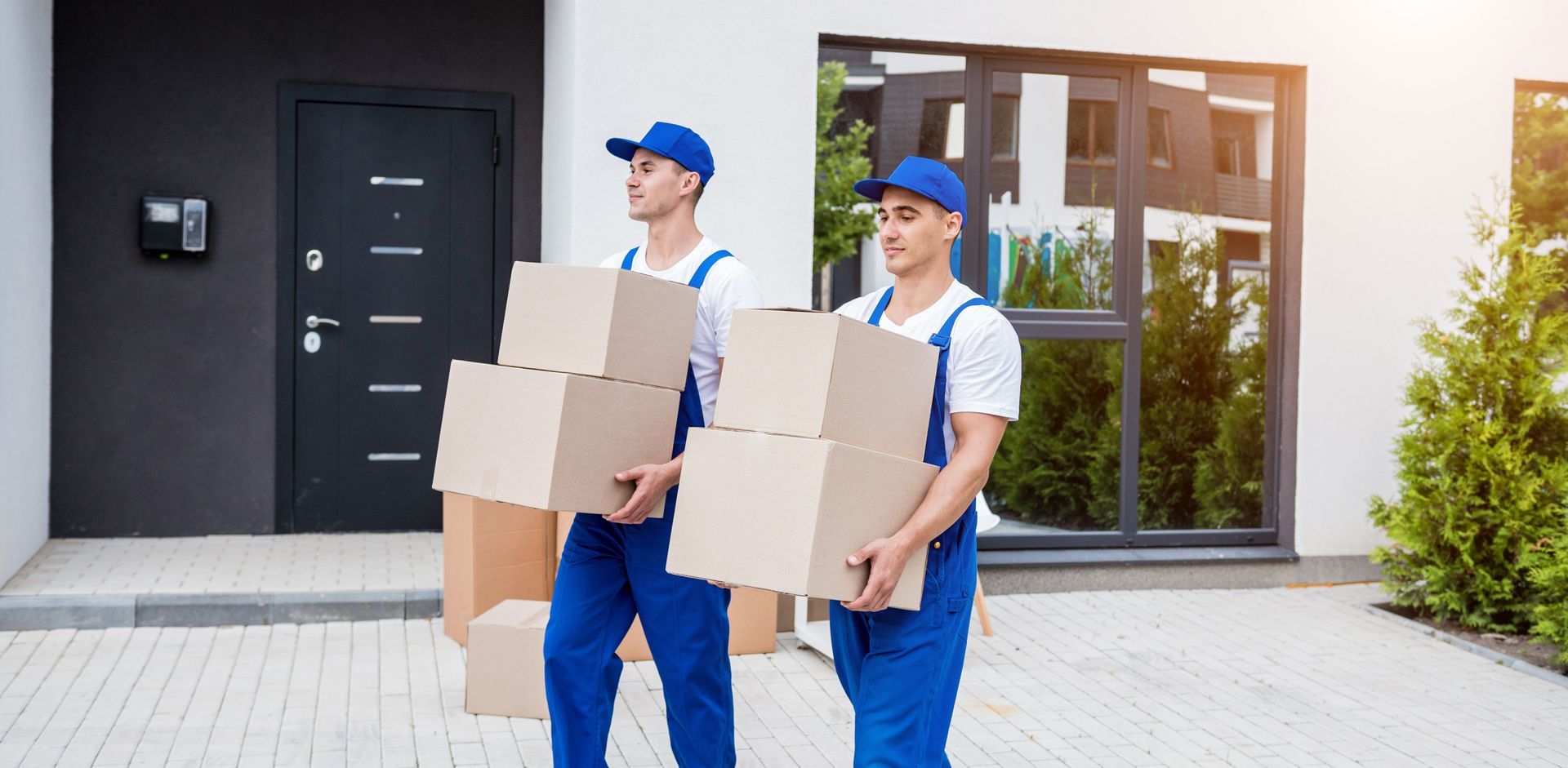 Two movers in blue uniforms carrying boxes outside a house.