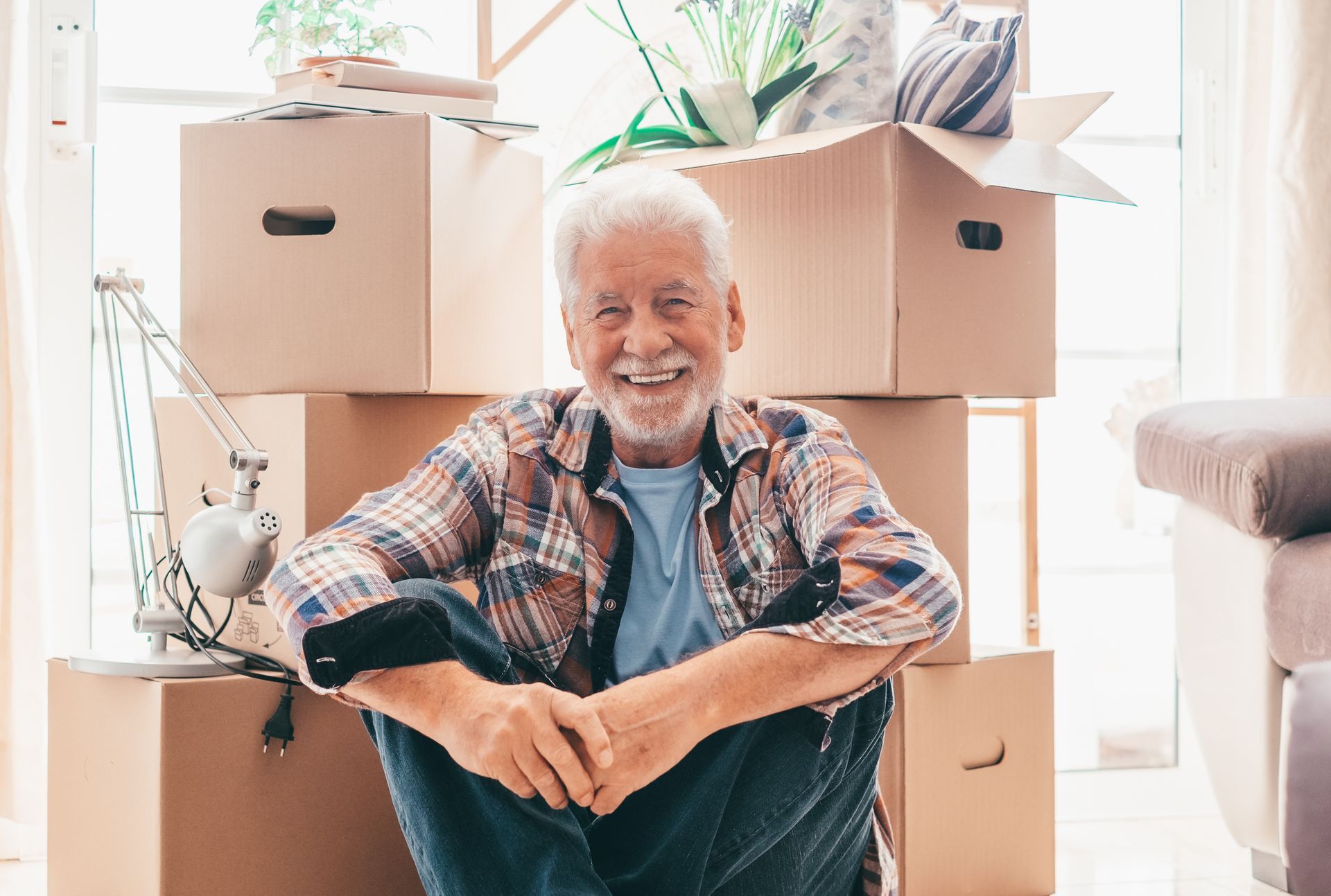 A smiling person sitting on the floor surrounded by cardboard moving boxes in a brightly lit room.