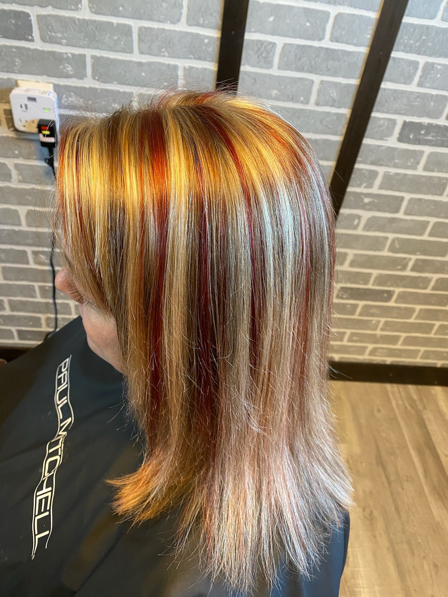 Woman with shoulder-length hair; red and blonde highlights. Sitting at a salon, brick wall background.