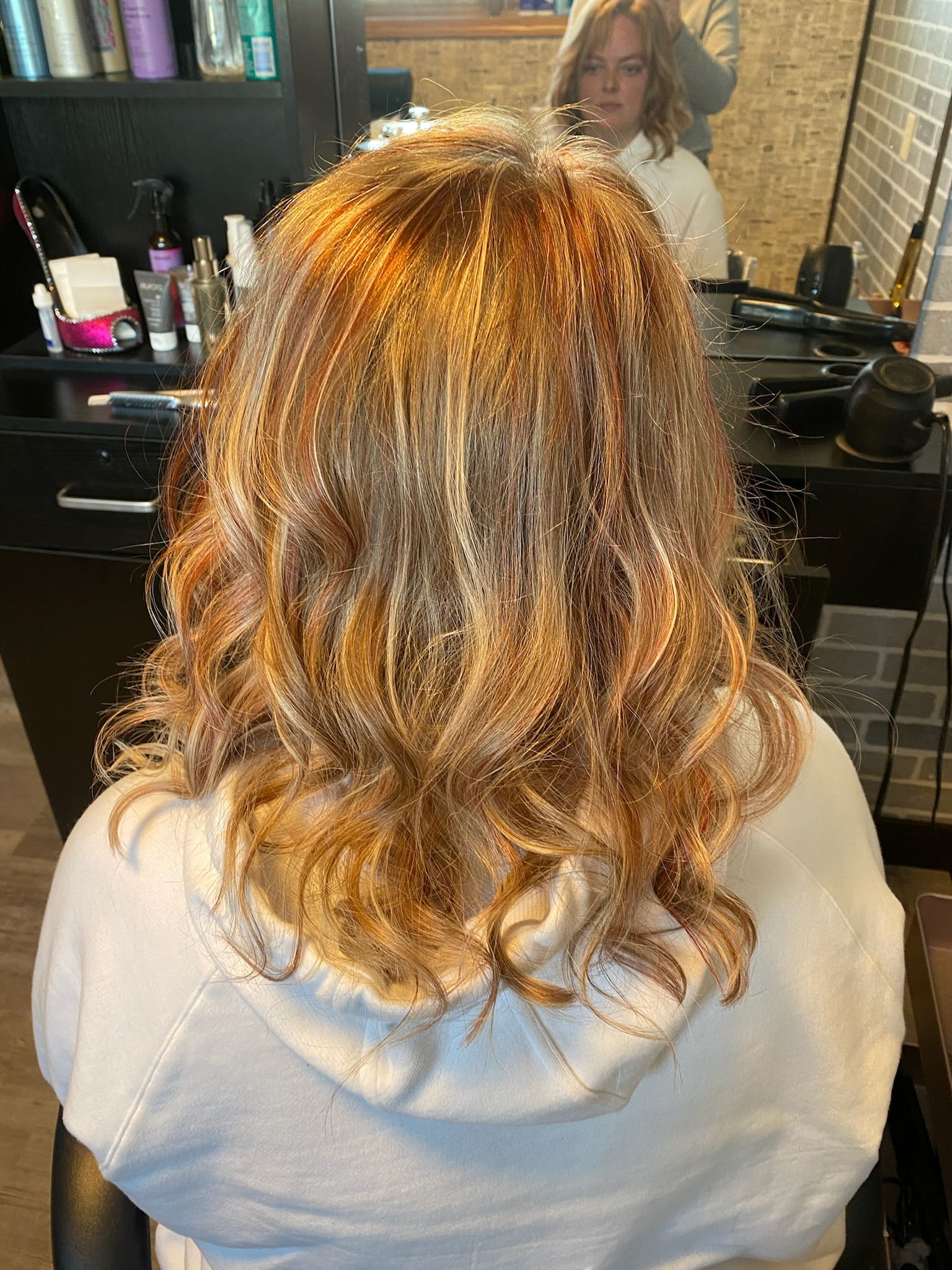 Person with highlighted, wavy hair in a salon chair; mirror reflection shows the stylist; warm tones.