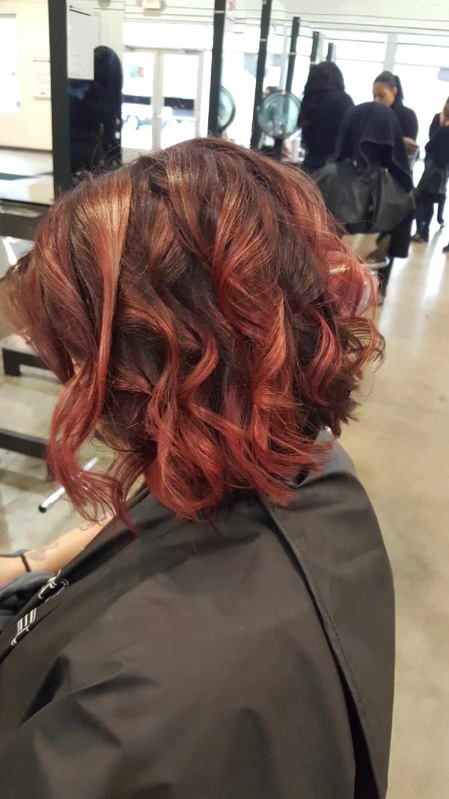 Woman with red-tinted, curly hair styled in a salon setting, wearing a black cape.