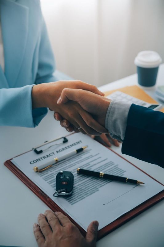 Two people shaking hands over a contract, car keys, and pens on a desk.