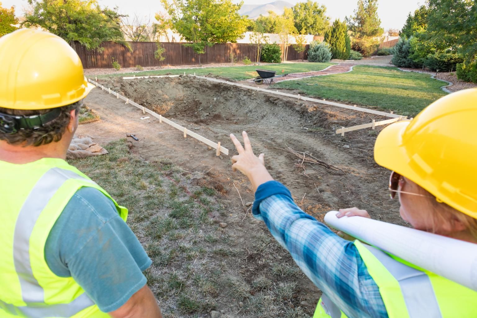 Two workers in safety gear and hard hats survey a backyard construction site where a trench has been dug for a project.