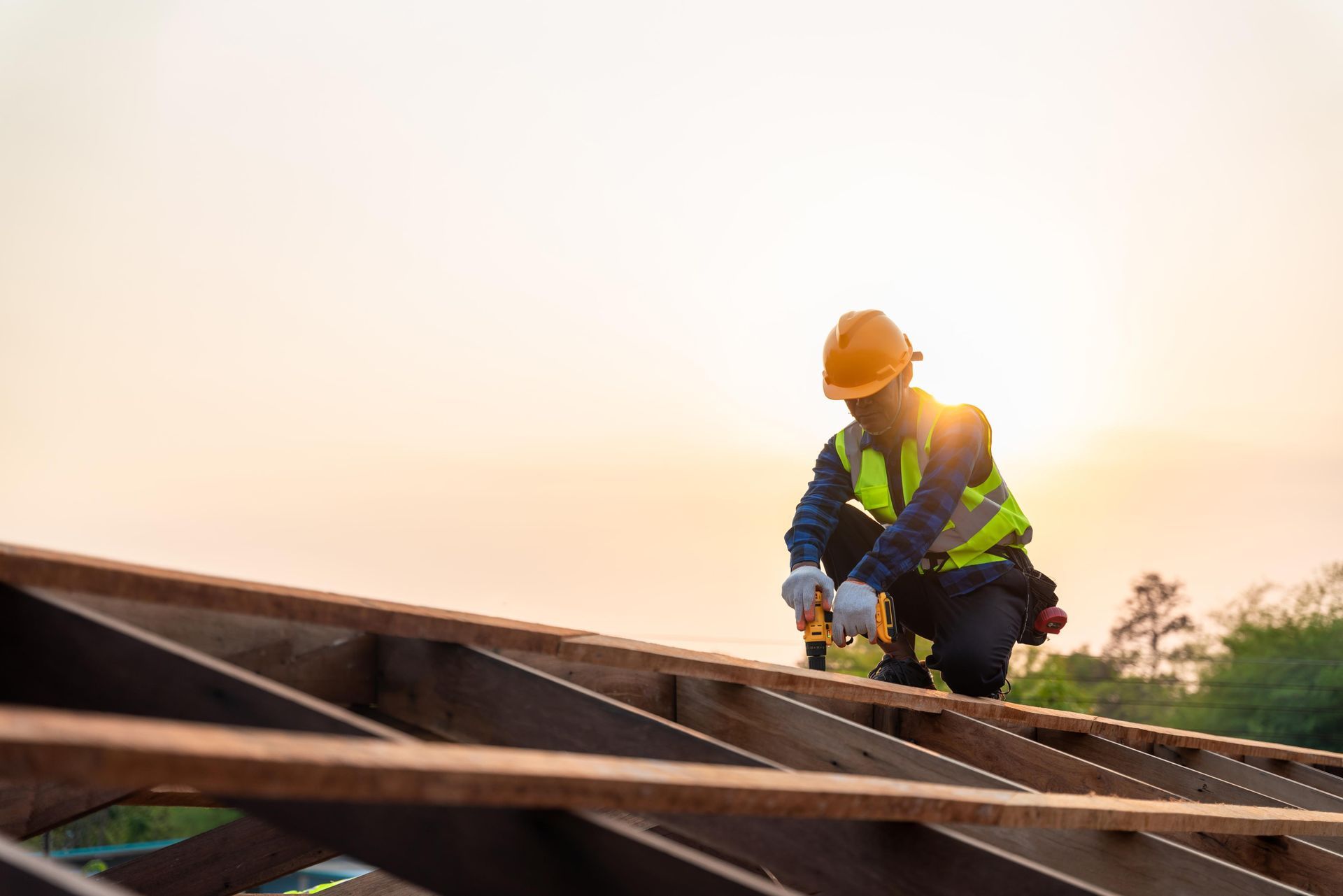A construction worker in a safety vest and hard hat works on a wooden roof frame at sunset.