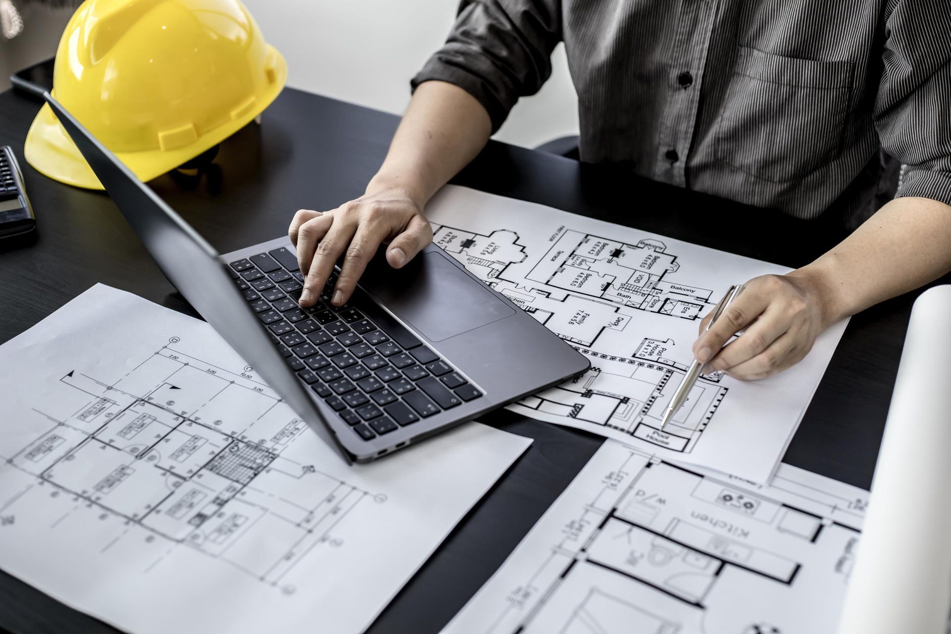 A person works on a laptop at a desk with architectural blueprints and a yellow construction hard hat.