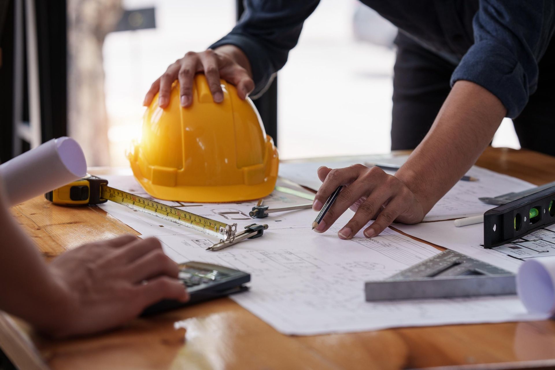 Two people review architectural blueprints on a wooden table with a yellow hard hat, tape measure, and drafting tools.