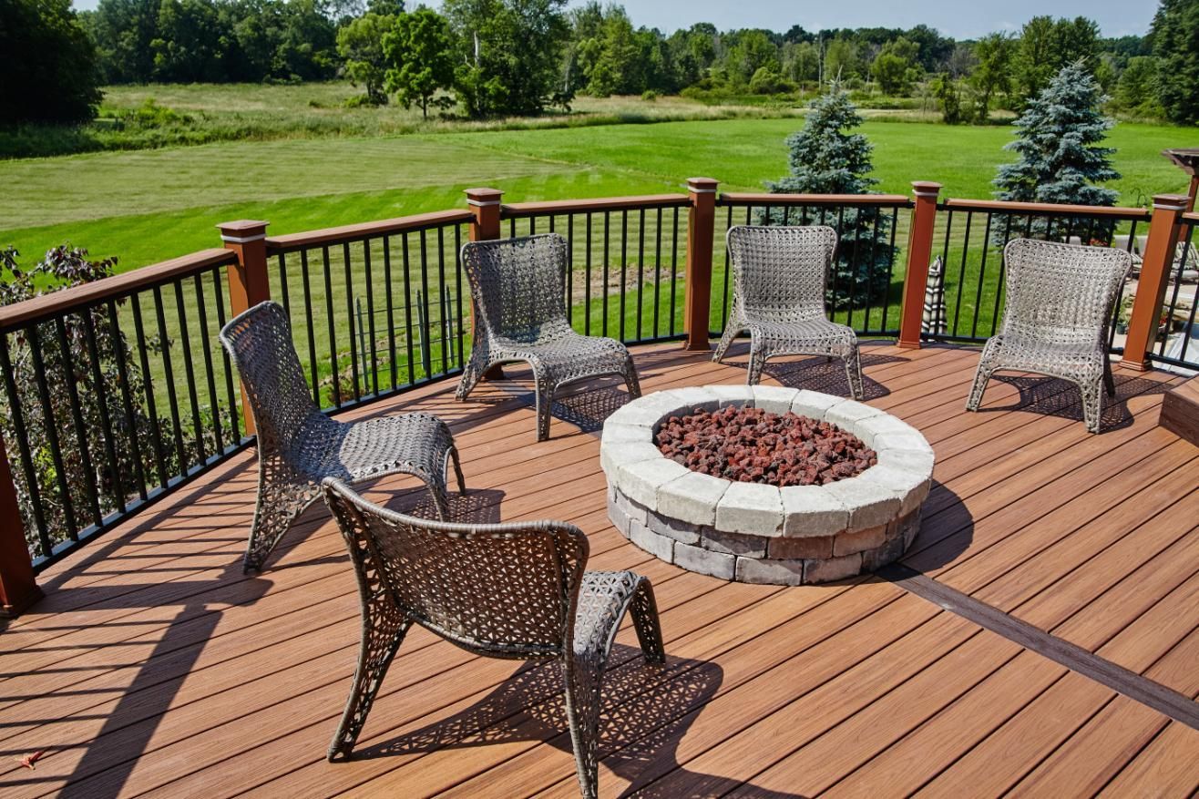 A circular stone fire pit sits on a wooden deck surrounded by four wicker chairs, overlooking a grassy meadow.