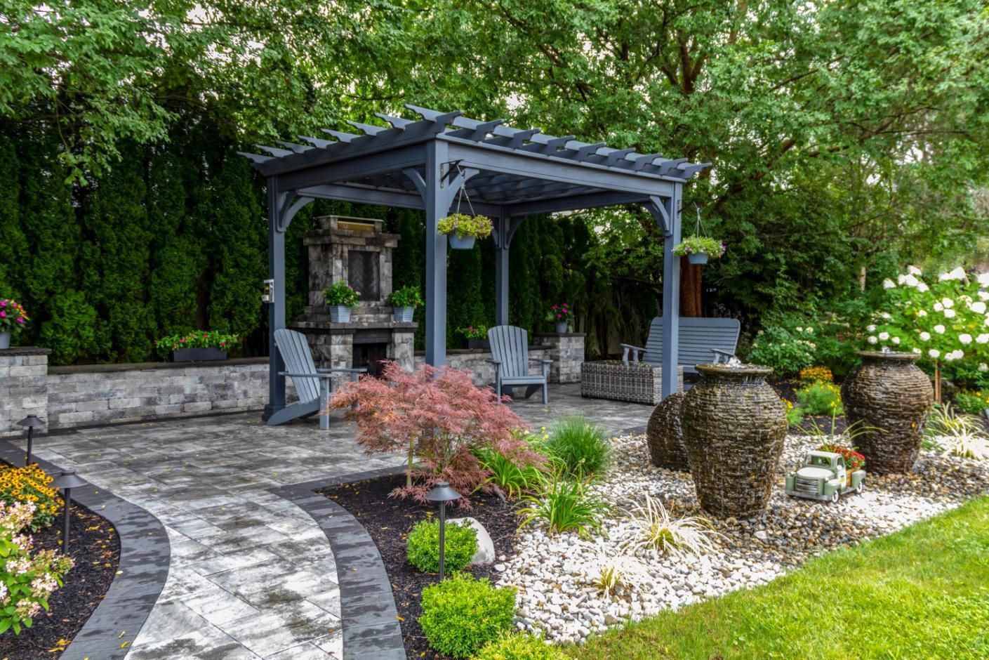 A backyard patio featuring a gray pergola, stone fireplace, outdoor seating, and decorative urns on a paved walkway.