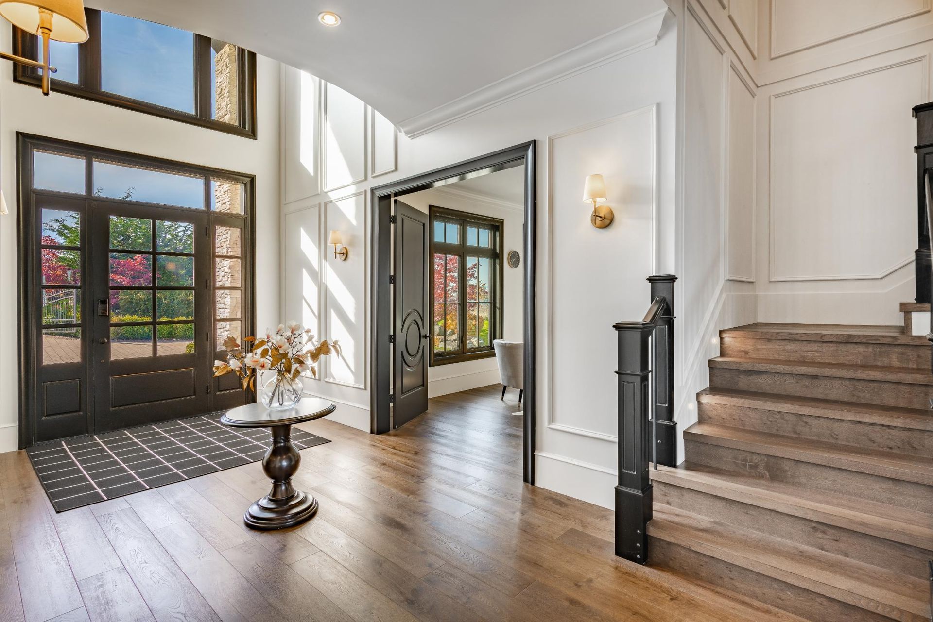 A bright, modern entryway with white walls, wood floors, a dark front door, a pedestal table, and a wooden staircase.