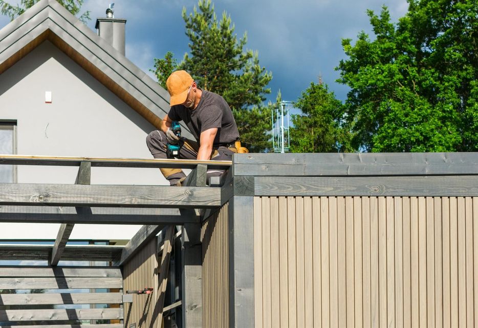 A person wearing a cap and work clothes uses a power drill to build a wooden patio structure outside a house.
