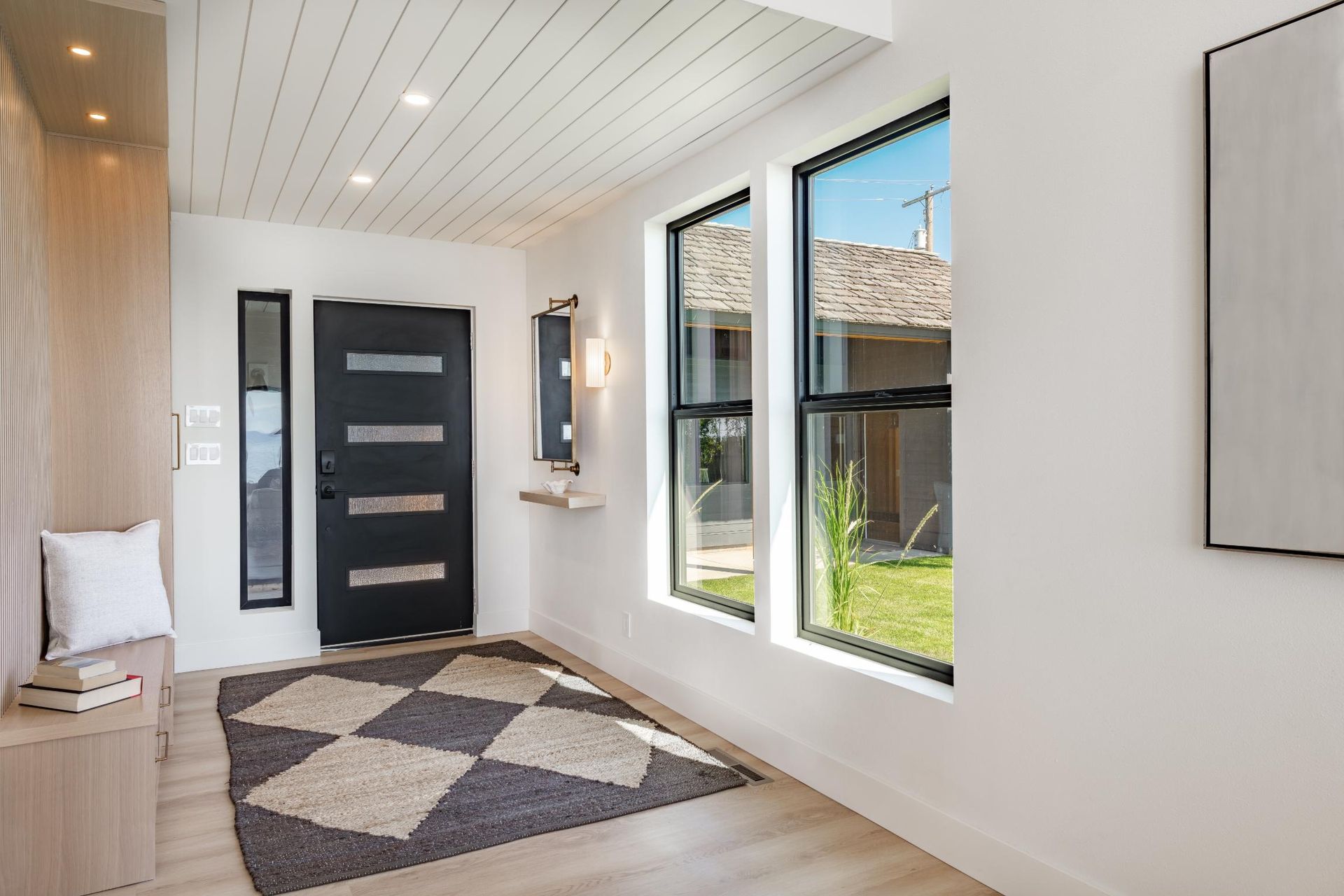 A modern entryway with a dark door, light wood walls, a geometric patterned rug, and large windows looking outside.