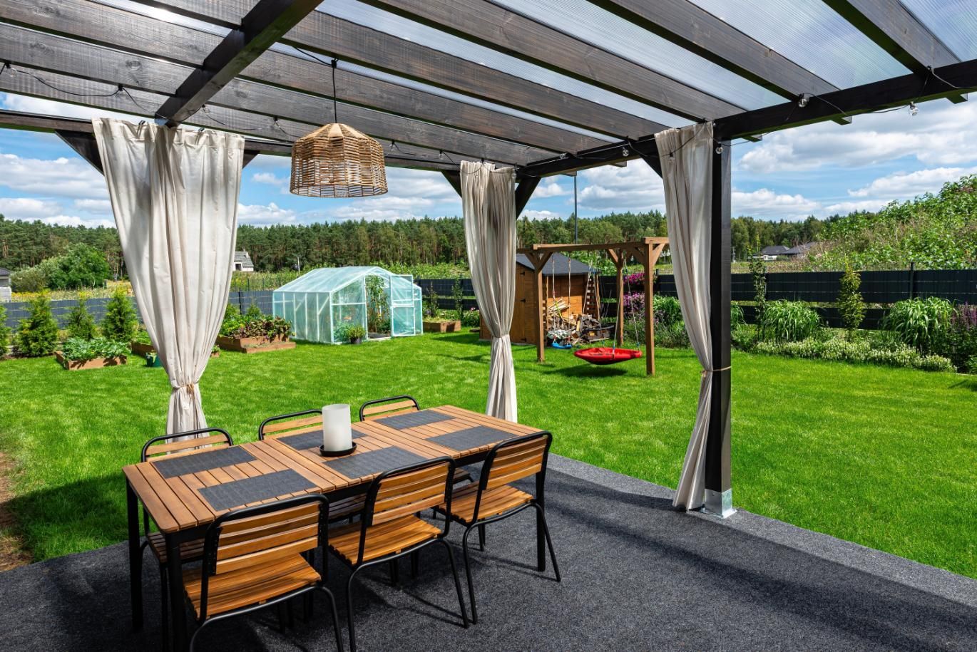 A dining set under a pergola on a patio, overlooking a green backyard with a greenhouse and a swing set.